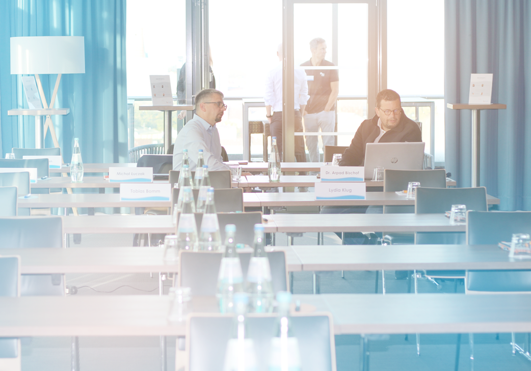 Conference room with several empty tables and chairs while two people sit at a table and work on a laptop